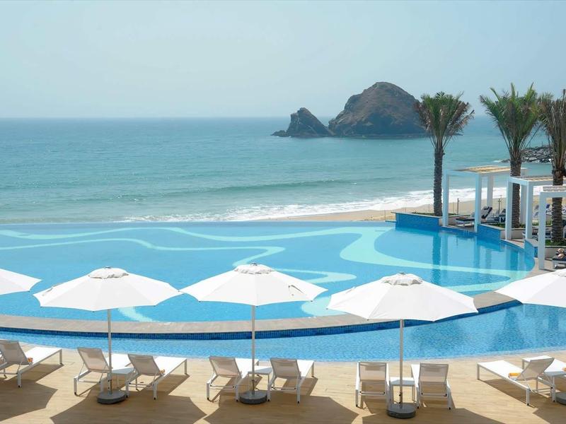 Infinity-Pool mit weißen Sonnenschirmen, Palmen und Meer im Hintergrund bei klarem Himmel.