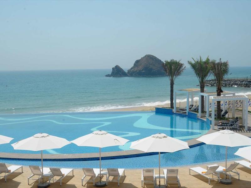 Hotelpool mit weißen Sonnenschirmen, Liegestühlen und Palmen vor blauem Meer und Felsen im Hintergrund.