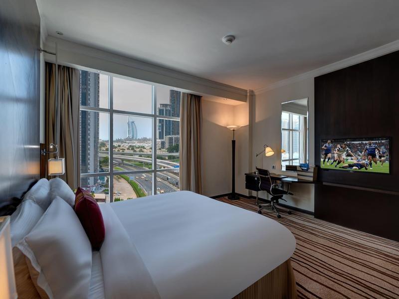 Modern hotel room with large bed, TV, desk, and city view through big windows.