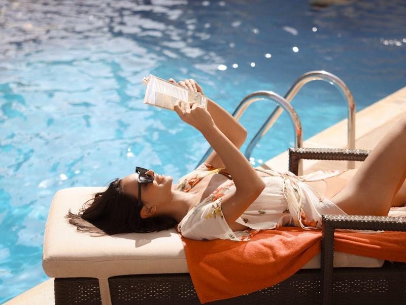 Woman relaxing on lounge chair by pool reading a book in sunshine.