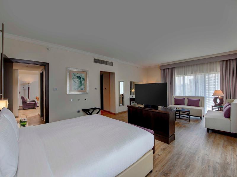 Modern hotel room with large bed, seating area, TV, and window with light curtains.