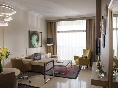 Habitación de hotel decorada modernamente con área de estar, escritorio y grandes ventanas con cortinas.