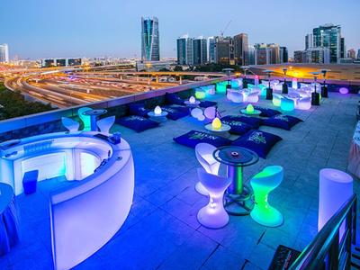 Terraza en la azotea con asientos iluminados en colores y vista a una ciudad moderna al atardecer.