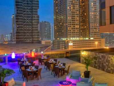 Terraza en la azotea con áreas de descanso y luces de colores frente a altos edificios al anochecer.
