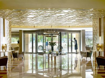 Luminosa recepción de hotel con candelabro moderno, puerta automática de vidrio y áreas de descanso a los lados.