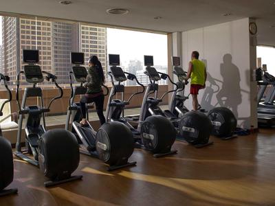 Gym with exercise machines and guests in a hotel room with large windows.