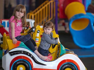 Dos niños sentados en un coche de juguete con un parque infantil interior colorido al fondo.