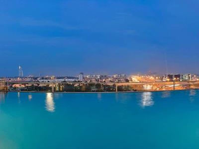Gran piscina iluminada con vista a la ciudad de noche, rodeada de edificios y reflejos en el agua.