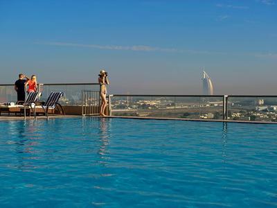 Piscina en la azotea con agua azul clara y personas junto a la barandilla, ciudad y Burj Al Arab al fondo.