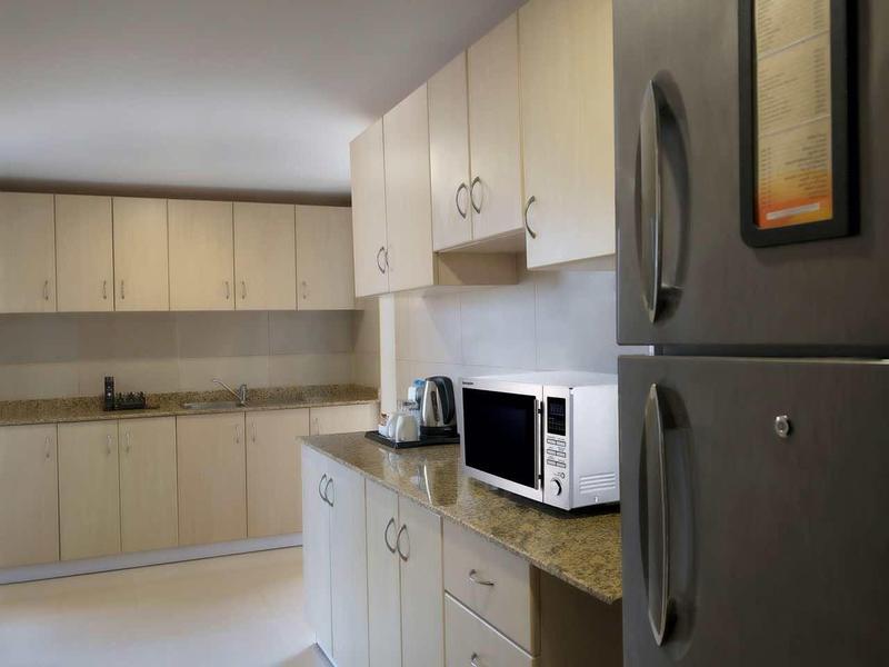 Área de cocina moderna con armarios claros, electrodomésticos y un refrigerador gris en una habitación de hotel.