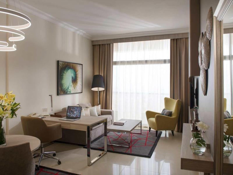 Habitación de hotel decorada modernamente con área de estar, escritorio y grandes ventanas con cortinas.