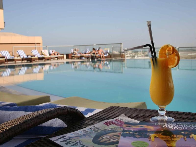 Vaso de cóctel sobre mesa junto a piscina con tumbonas en azotea de hotel bajo cielo despejado.
