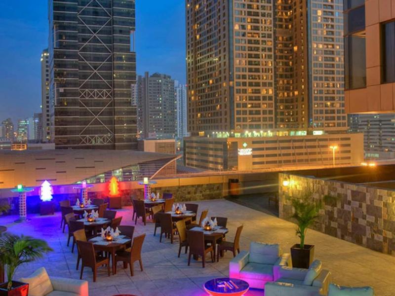 Terraza en la azotea con áreas de descanso y luces de colores frente a altos edificios al anochecer.