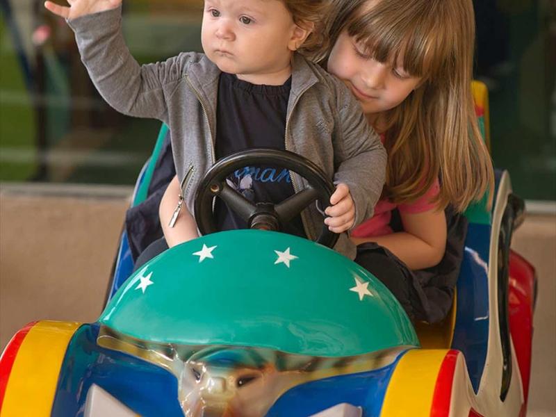 Niño pequeño al volante de un vehículo colorido, niña lo observa atentamente.