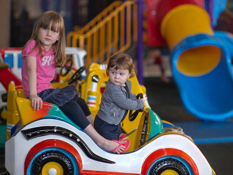 Dos niños sentados en un coche de juguete con un parque infantil interior colorido al fondo.
