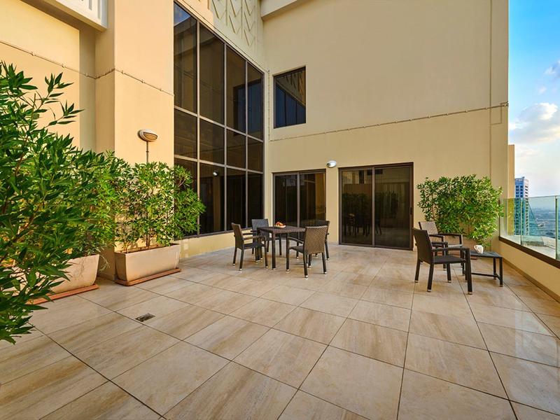 Terras met tafels en stoelen, omgeven door planten en hoge gebouwen op de achtergrond.