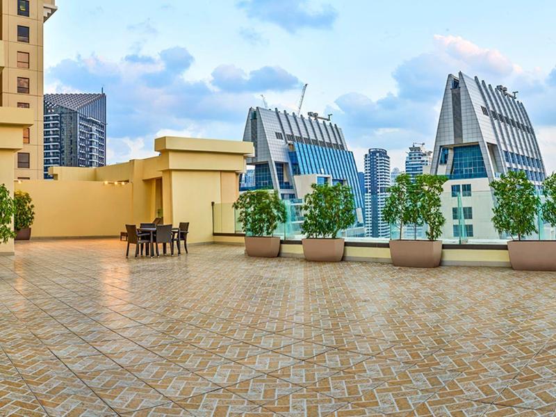 Groot terras met tafels, stoelen en bloempotten voor moderne stadsgebouwen onder een bewolkte hemel.