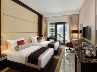 Modern hotel room with two single beds, desk, TV, and large windows overlooking the city.