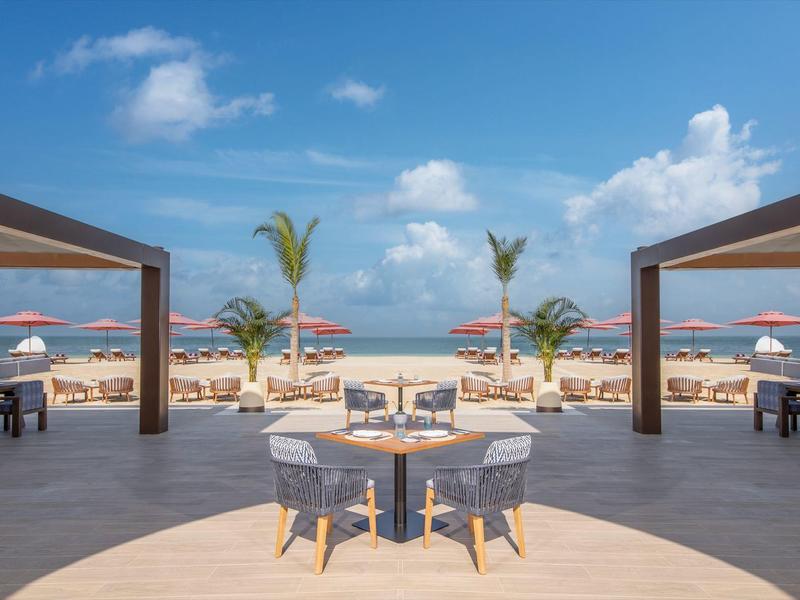 Moderner Außenbereich eines Hotels am Strand mit Tischen, Stühlen und Sonnenschirmen.