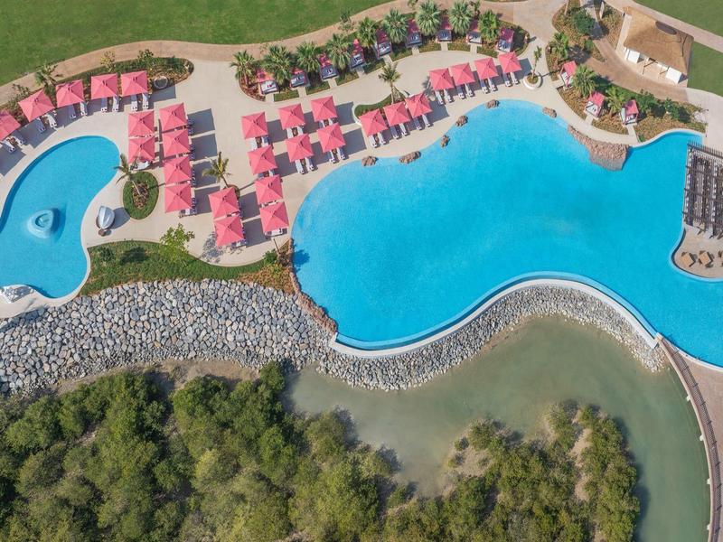 Luftaufnahme eines Hotels mit zwei großen blauen Pools, Sonnenschirmen und Liegestühlen am grünen Gelände.