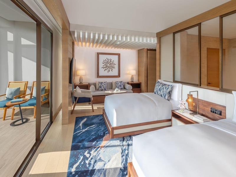 Helles Hotelzimmer mit zwei Betten, blauen Teppichen und Zugang zu einer Terrasse mit Sitzecke.