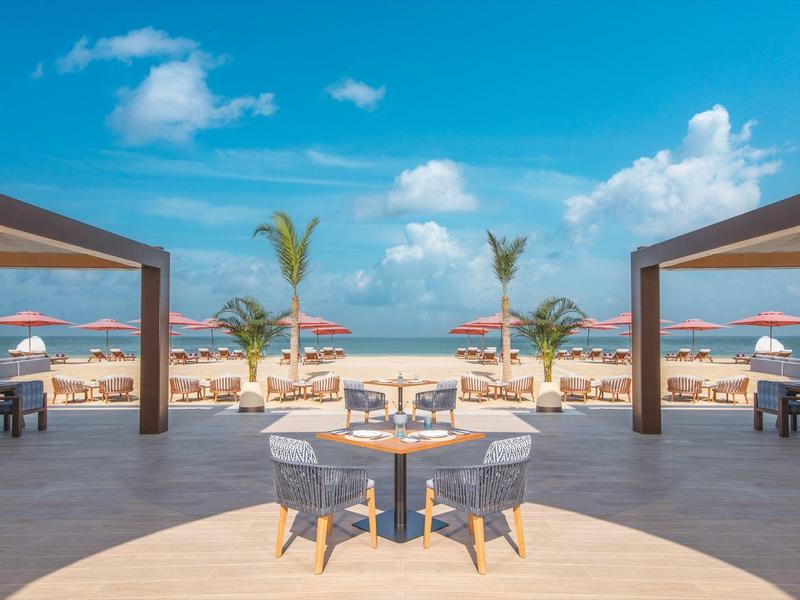 Offenes Strandrestaurant mit Tischen und Sonnenschirmen am Meer unter blauem Himmel.