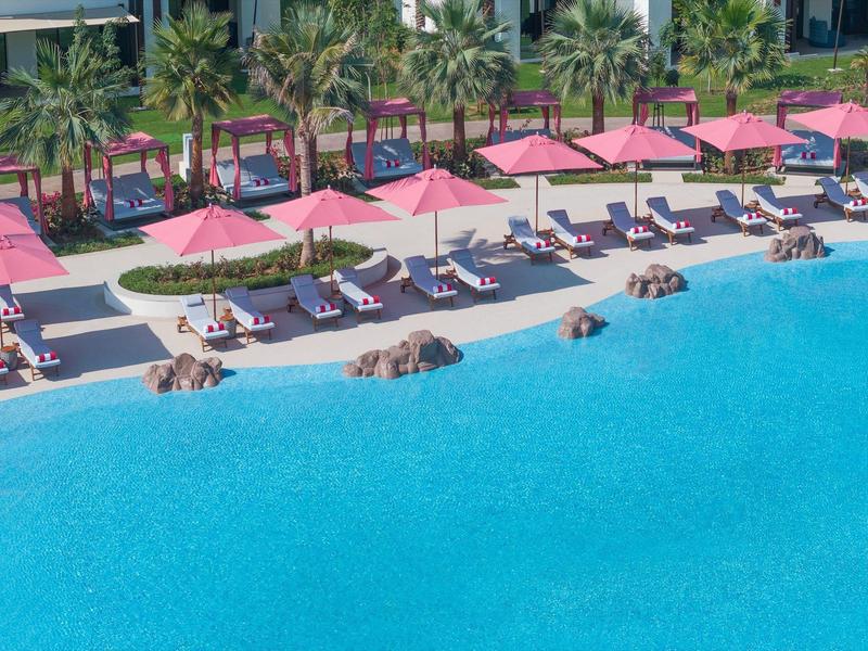 Großer Pool mit blauer Wasserfläche und Sonnenliegen unter pinken Sonnenschirmen neben einem Hotel.