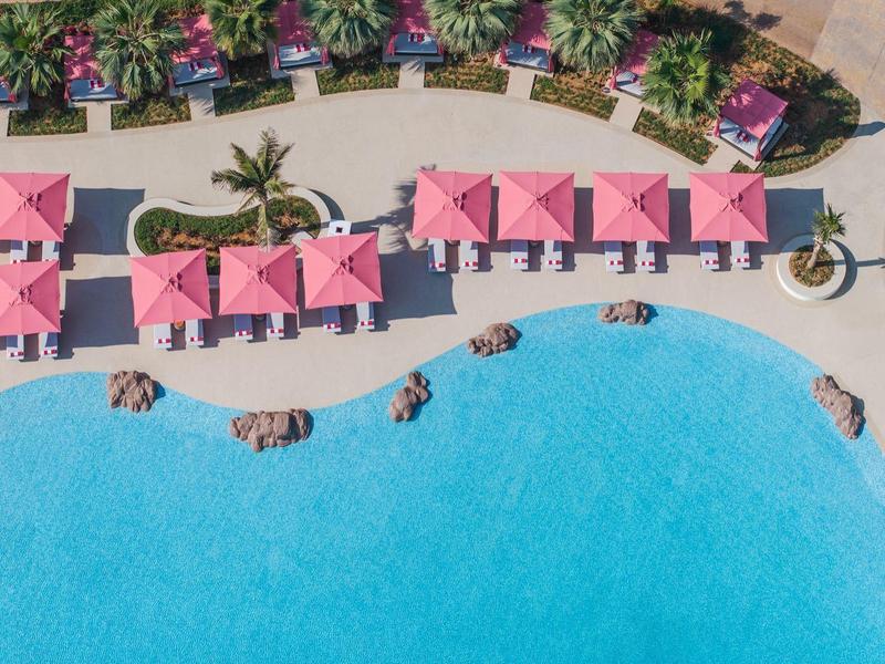 Vogelperspektive eines blauen Pools mit rosa Schirmen und Liegestühlen in einem Hotelgarten.