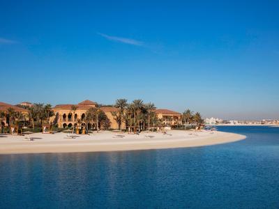 Strand mit weißem Sand, blauem Wasser und Gebäuden im Hintergrund bei klarem Himmel.