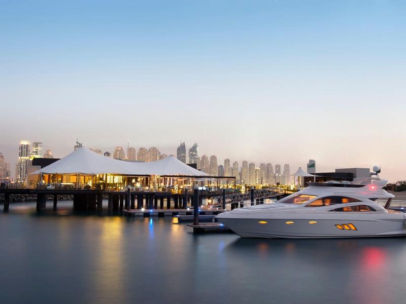 Luxusyacht liegt im Hafen neben einer modernen Marina mit Gebäuden im Hintergrund bei Sonnenuntergang.