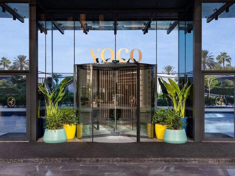 Eingang mit Glasdrehtür, Palmen in gelben und blauen Töpfen, reflektierende Glasfassade.