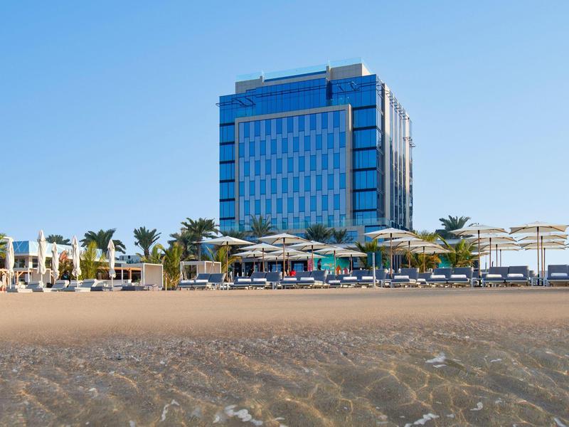 Modernes Hotelgebäude am Strand mit Liegestühlen, Palmen und klarem blauem Himmel.
