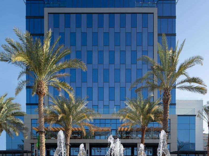 Blauer Glasbau mit fünf Palmen und mehreren Wasserfontänen vor klarem Himmel.