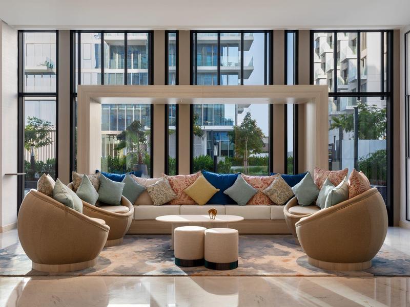 Helles Wohnzimmer mit großen Fenstern, runder Couch, Sesseln und zwei kleinen Hockern auf Teppich.