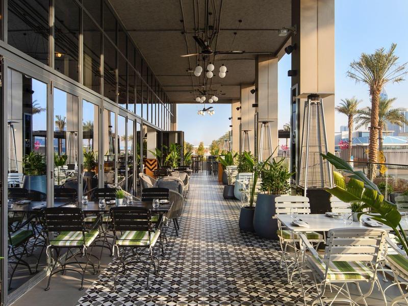 Offene Café-Terrasse mit schwarz-weißem Fliesenboden, grünen Stühlen und Pflanzen entlang der Seite.