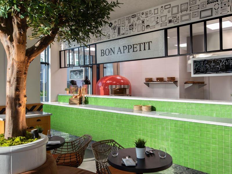 Gemütliches Café mit grünem Tresen, Baum, runden Tischen und rotem Pizzaofen im Hintergrund.