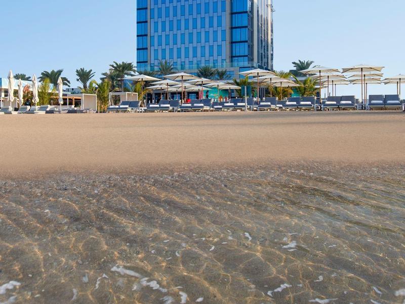 Klares Meer mit Wellen vor einem modernen Gebäude und Palmen unter blauem Himmel am Strand.