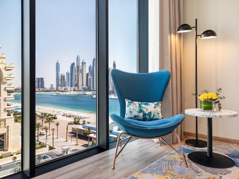 Gemütlicher blauer Sessel mit Kissen neben rundem Tisch und Stehlampe vor großem Panoramafenster.