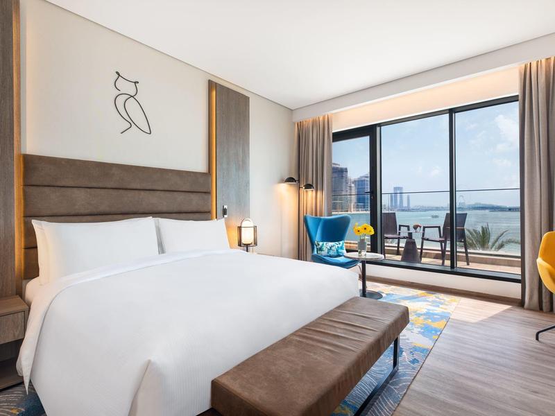 Helles Hotelzimmer mit großem Bett, Sitzbank, modernem Stuhl und Balkon mit Aussicht auf Wasser.