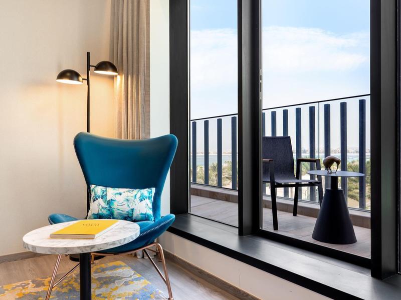 Moderner Lesesessel mit blauem Stoff und buntem Kissen neben rundem weißen Tisch am Fenster mit Balkonblick.