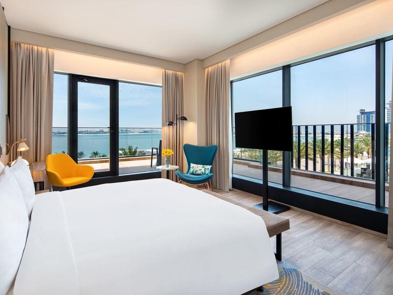 Modernes Hotelzimmer mit großem Bett, Stühlen, zwei Fenstern und Balkon mit Meerblick.