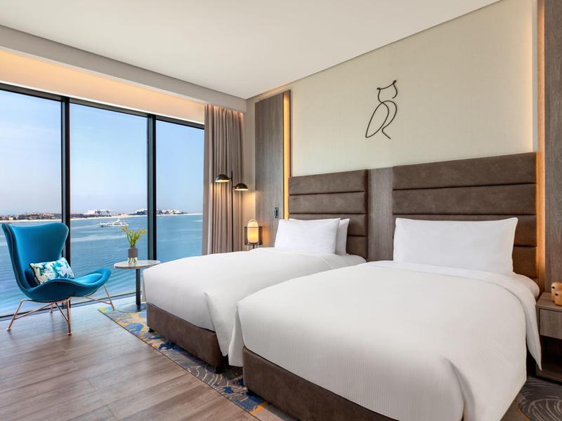 Modernes Hotelzimmer mit zwei weißen Betten, blauer Sitzgruppe und großem Fenster mit Meerblick.