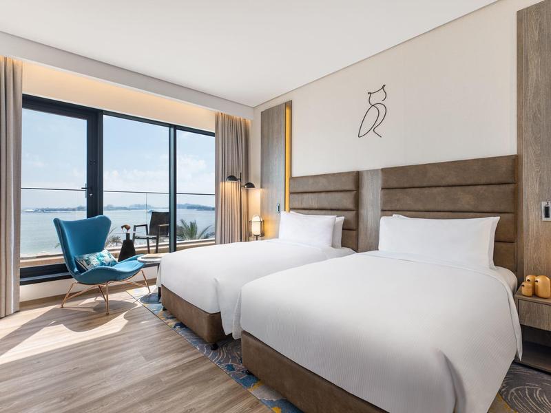 Elegantes Hotelzimmer mit zwei weißen Betten, blauem Sessel und großem Fenster mit Meerblick.