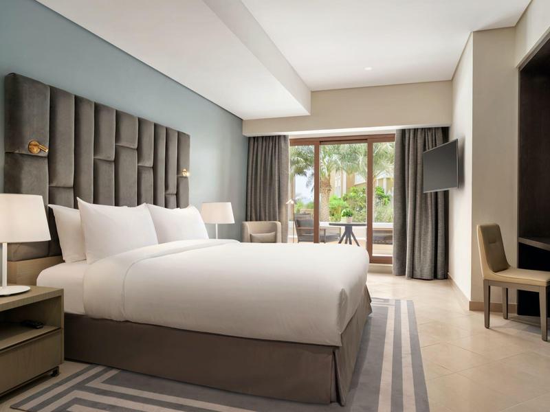 Modern eingerichtetes Hotelzimmer mit großem Bett, Nachttischen, Lampen und Blick auf Balkon.