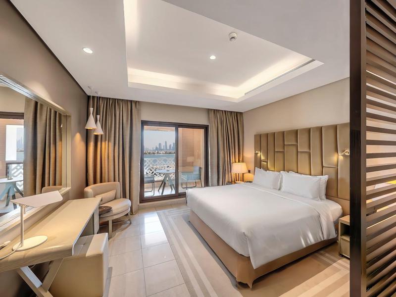 Modernes Hotelzimmer mit großem Bett, Schreibtisch, Stuhl und Balkon mit Blick auf das Wasser.