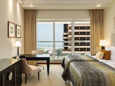 Hotelkamer met twee bedden, bureau, fauteuil, lampen en balkon met uitzicht op gebouwen en zee.