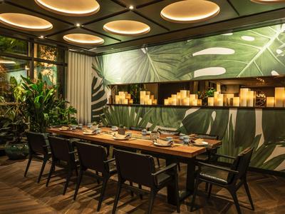 Moderne eetkamer met houten tafel, zwarte stoelen, groen bladbehang en plafondlampen.