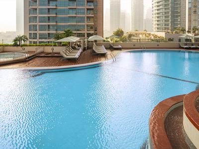 Duży basen na zewnątrz z leżakami i nowoczesnymi wieżowcami w tle.