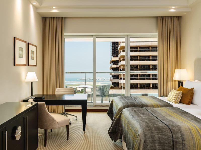 Hotelkamer met twee bedden, bureau, fauteuil, lampen en balkon met uitzicht op gebouwen en zee.