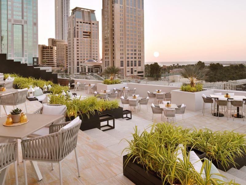 Moderne buitenruimte met tafels, stoelen, planten en stadsgebouwen bij zonsondergang.